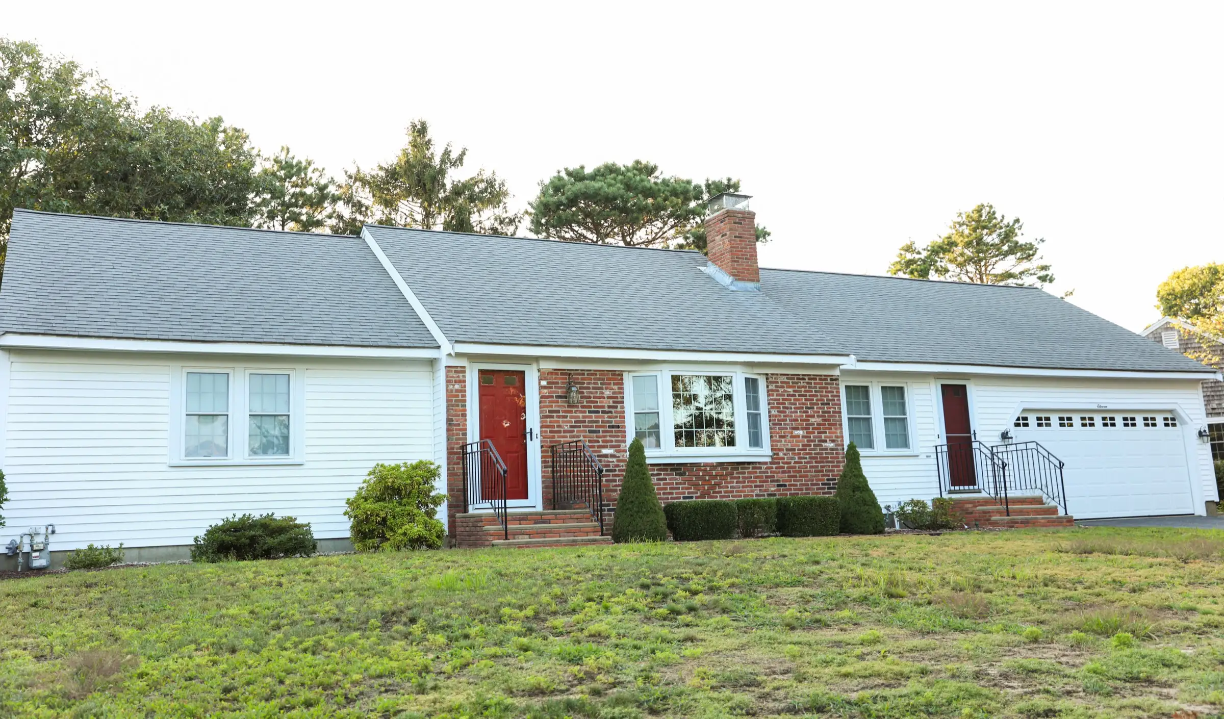 White and brick ranch house with a patchy lawn and pine trees behind